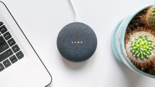 Google home mini smart speaker on a desk between a MacBook and a cactus