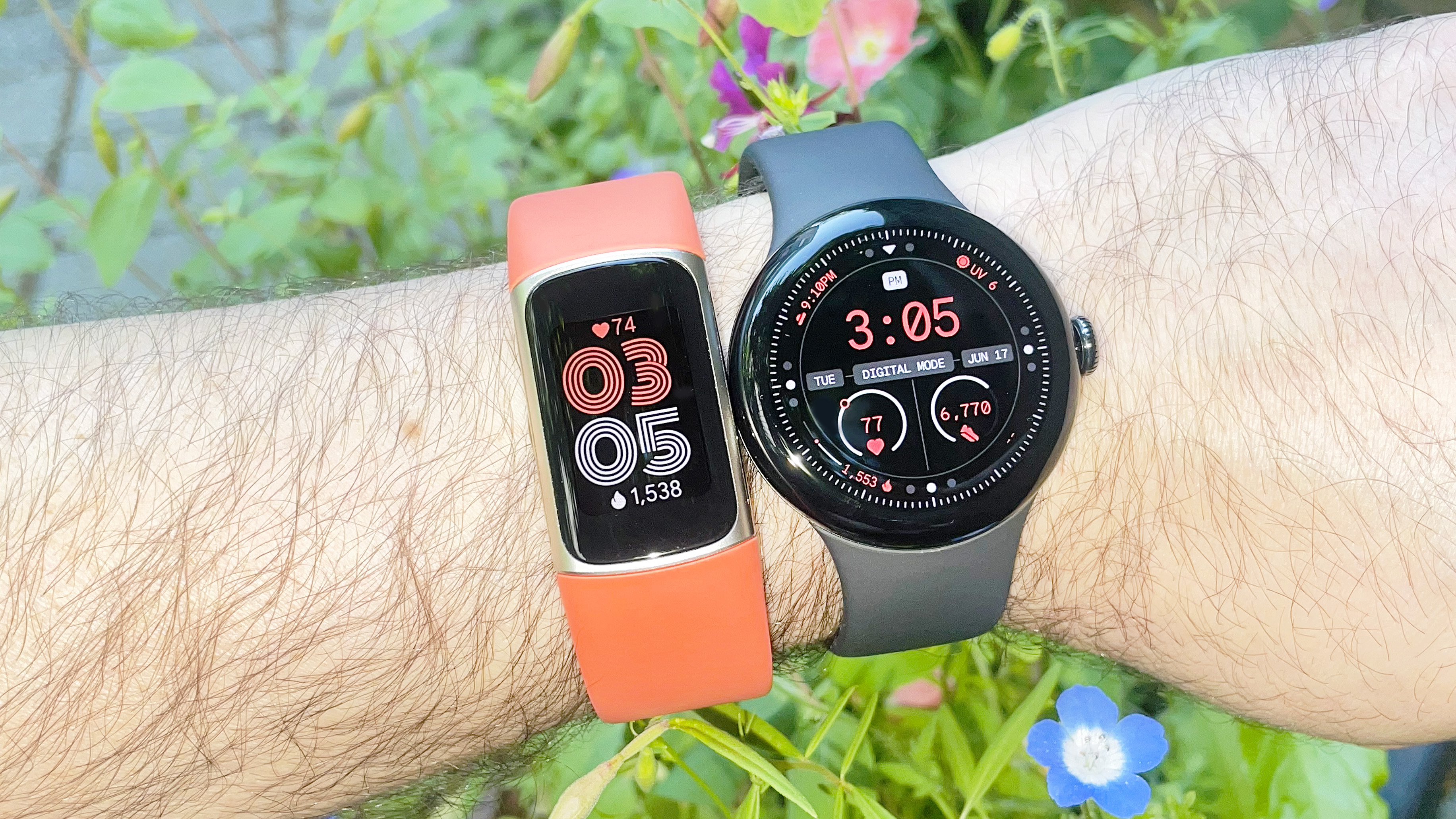 The Fitbit Charge 6 on an orange strap next to the Pixel Watch 3 on a black strap on a user&#039;s wrist with blue and pink flowers in the background