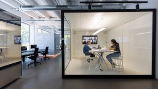 Workplace monitoring concept image showing office workers sitting at desk during video call in a glass box.