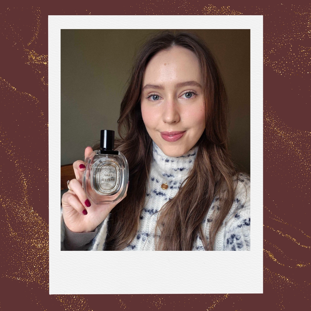 A picture of Digital Beauty Writer, Naomi Jamieson, seen wearing a black and white speckled jumper and holding a bottle of Diptyque's L'eau Papier/ in a burgundy template with a gold glitter overlay