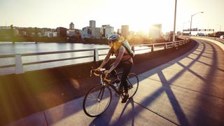 Woman cycling down a river pathway in an industrial city, wearing jeans, backpack and a bike helmet