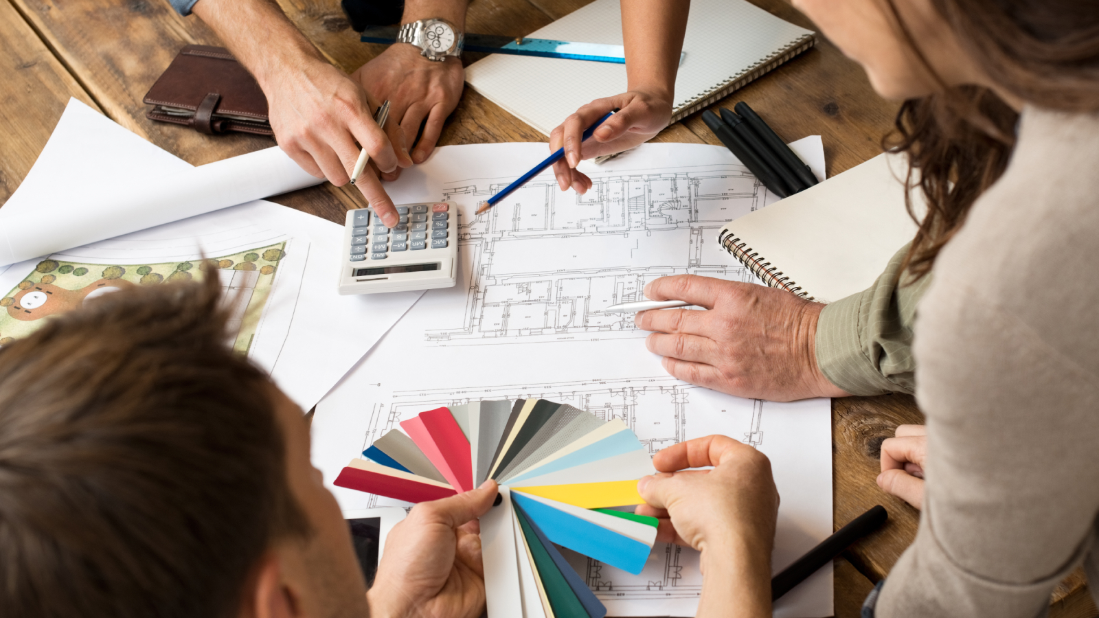 architect plans on table with people stood around holding various items including colour charts, calculator and pencils