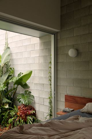 Image of a bedroom where the walls are made out of concrete blocks. There is one wall open to an outdoor area filled with plants. The bed frame is low and made out of wood.