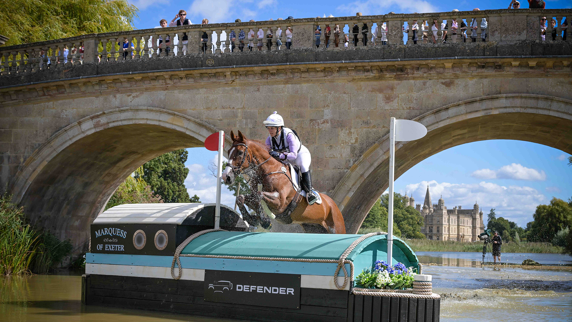 Gemma Stevens, a rider wearing a white hat cover and a pale purple top, rides chestnut horse Chilli Knight over a boat cross-country fence in water in front of a bridge at Burghley 2026