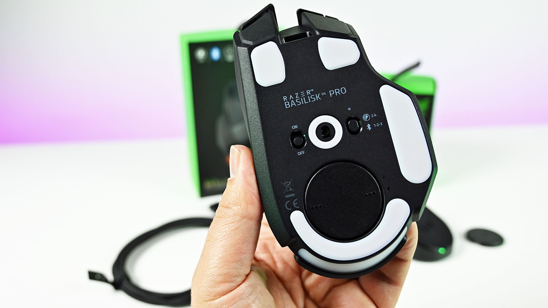 Underside view of the Razer Basilisk V4 Pro held in hand, showing white PTFE feet, optical sensor, profile button, and connection mode switch.