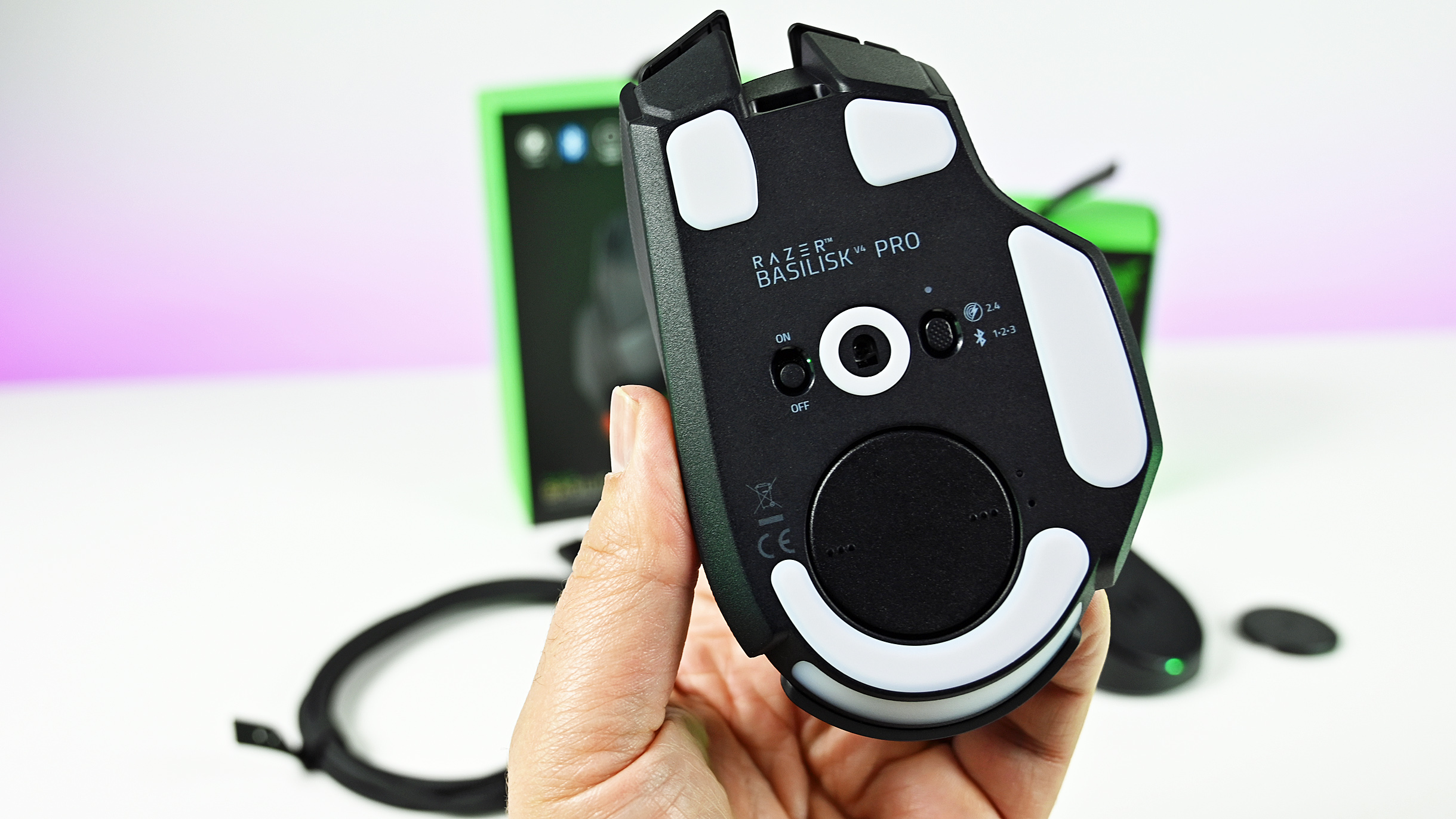 Underside view of the Razer Basilisk V4 Pro held in hand, showing white PTFE feet, optical sensor, profile button, and connection mode switch.