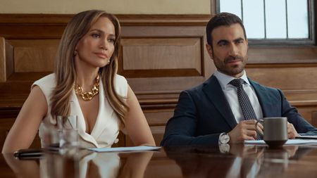 From left to right: Jennifer Lopez and Brett Goldstein sitting at a table side by side in Office Romance. 