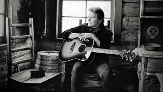 John Fogerty in a log cabin with an acoustic guitar 