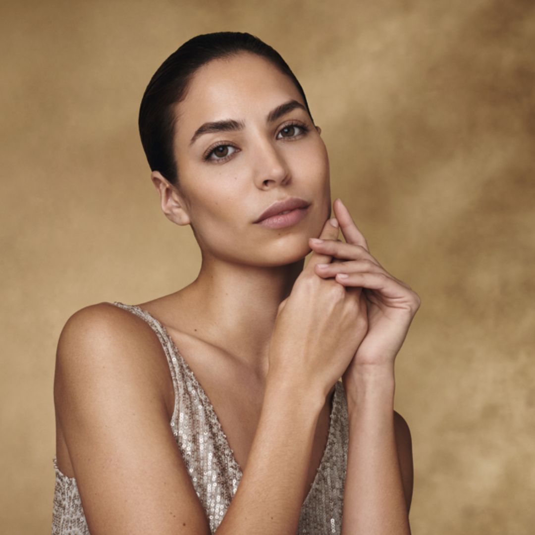 A woman with dark hair in a ponytail and smooth skin pictured in a golden room in a sequinned dress