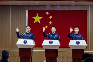 Three men in blue flight suits wave from podiums with the red Chinese flag behind them.