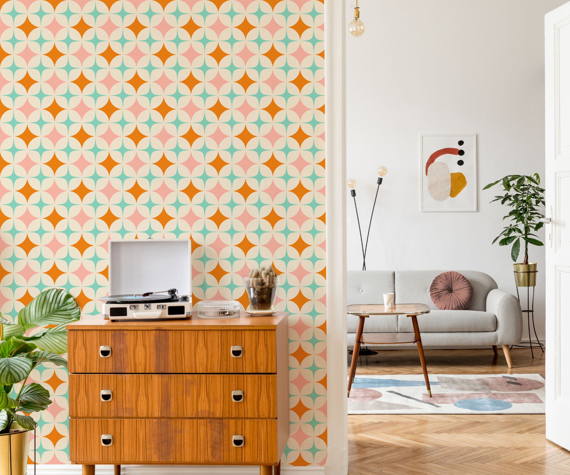 Retro hallway with bold geometric wallpaper in pink, orange and mint. A wooden chest with a record player and plants sits against the wall, leading into a pale lounge with a grey sofa.