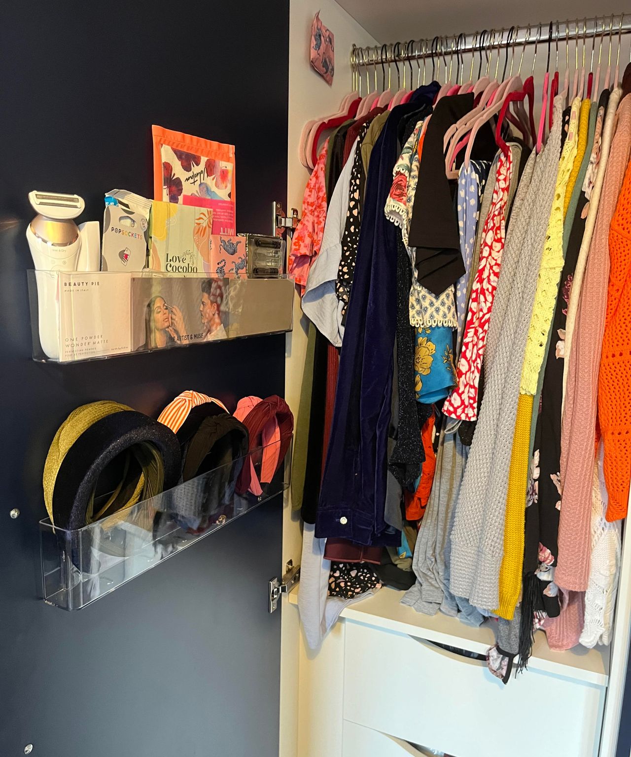 Viewing at an angle my clear plastic adhesive organizers inside my matte dark blue closet which is open and shows on the right a hanging rail of clothes with white drawers under