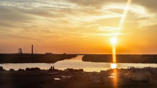 A sunrise streaks golden light out over the water near a silhouette of a rocket on a launch pad on the left of the image