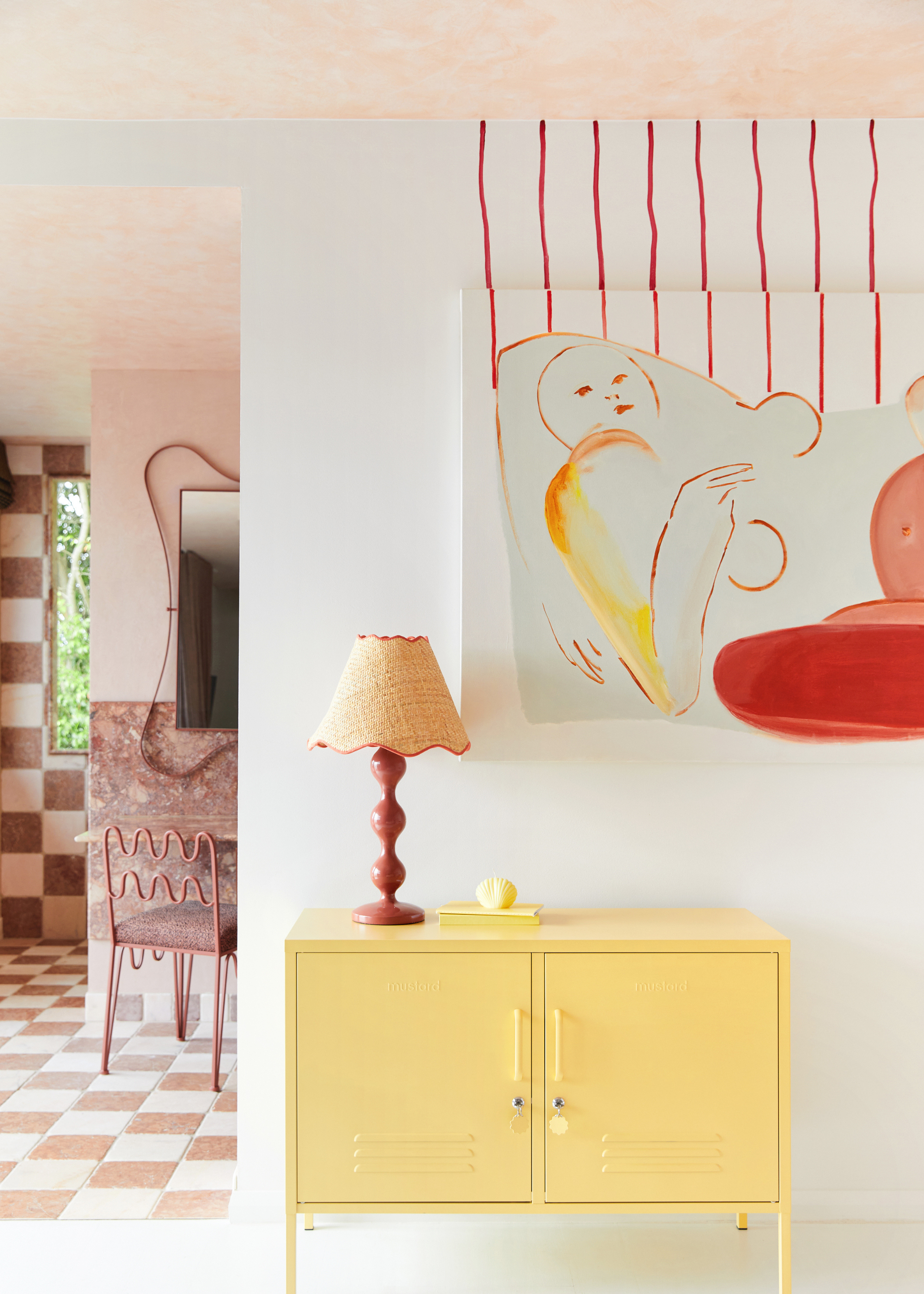 A white living room with a yellow locker-style cabinet, a curved burgundy lamp with a woven shade and a mural on the walls. Beside is a doorway to a room with checkerboard tiles, a metal seat and a metal frame around an art piece