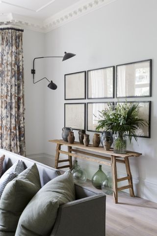 pale blue living room with lime washed floor boards, gray sofa, print drapes, bench with jugs, gallery wall and wall light