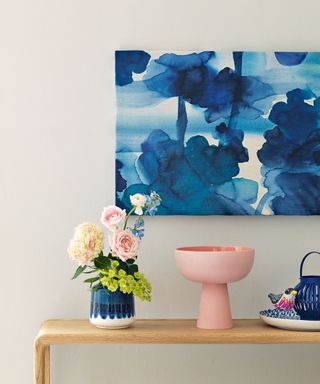 Wooden console table below a blue and white picture on a grey wall, blue and white vase with flowers and pink dish.