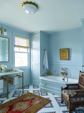 A light blue bathroom painted in Benjamin Moore's Yarmouth blue with a red/brown rug and wooden armchair next to the bathtub.