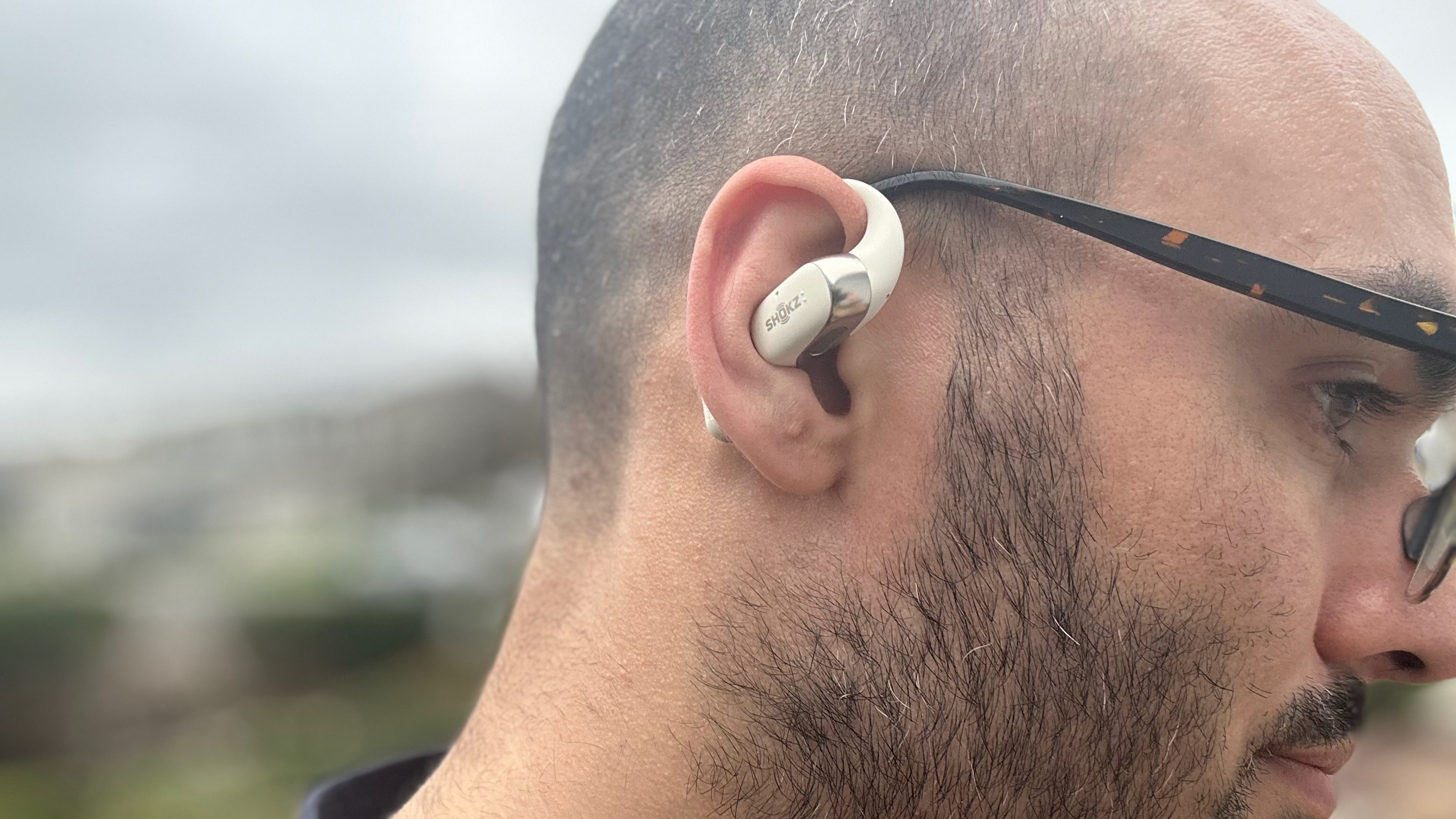 A photo of the reviewer wearing the Shokz OpenFit 2, with its ear hook looped next to his glasses.