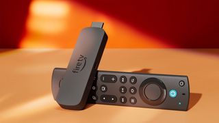 A stick-like device with a HDMI port sticking out the top of it is leaning on a black remote controller, all on an orange surface and background.