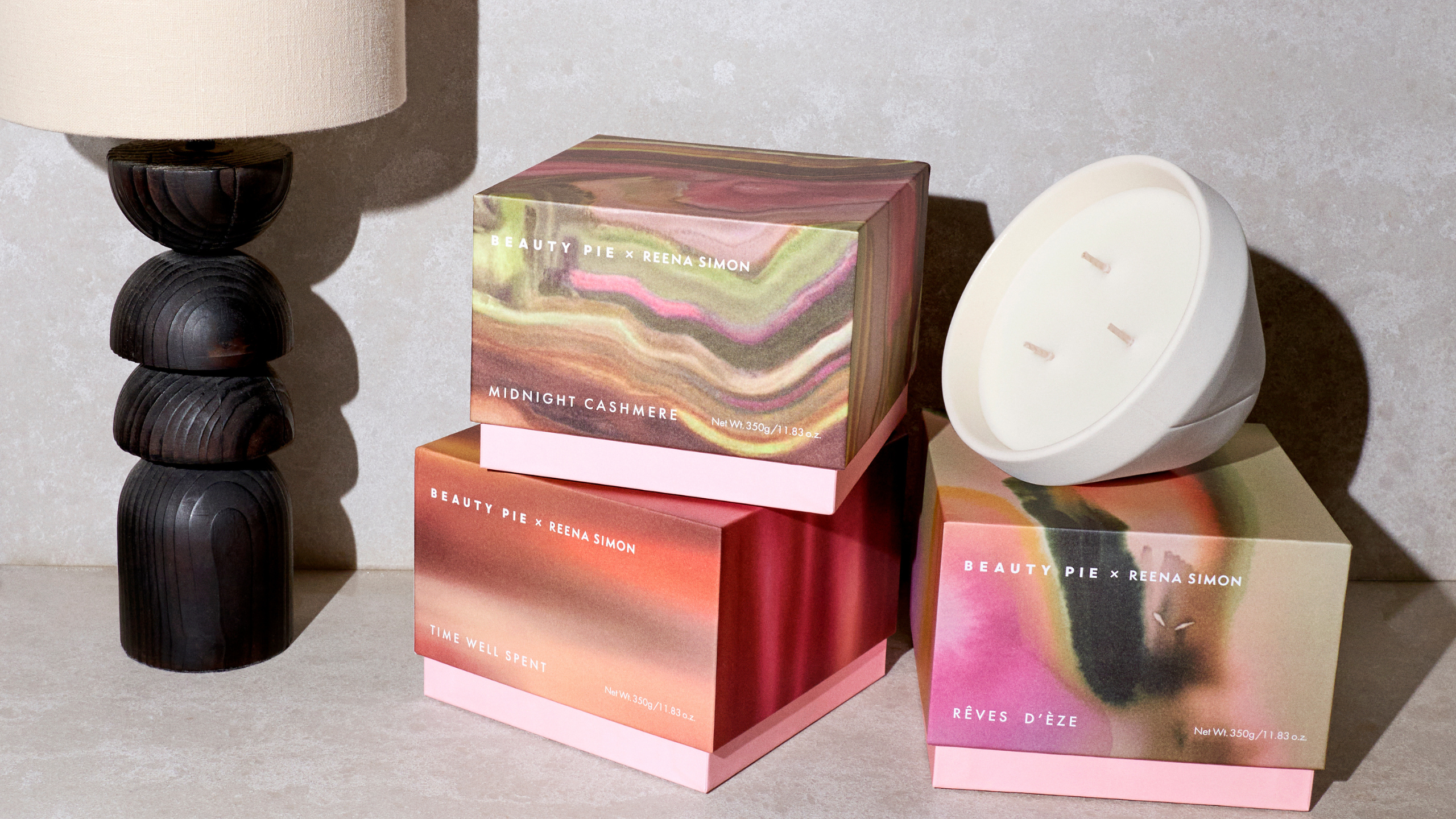 A stone surface with three colorful boxes and a tilted ceramic three-wick candle beside a structured lamp