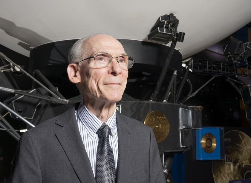 a smiling older man in a suit stands in front of a model of a large spacecraft. 