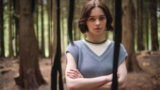 Emma Myers in a blue jumper and white shirt stands in the woods as Pip in A Good Girl's Guide to Murder.
