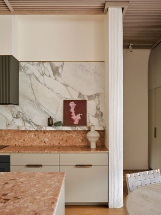 A beige and light pink kitchen with marble-blocked walls and a terracotta-colored countertop and backsplash. There is a cream vase on the counter and a plum painting on the marble shelf