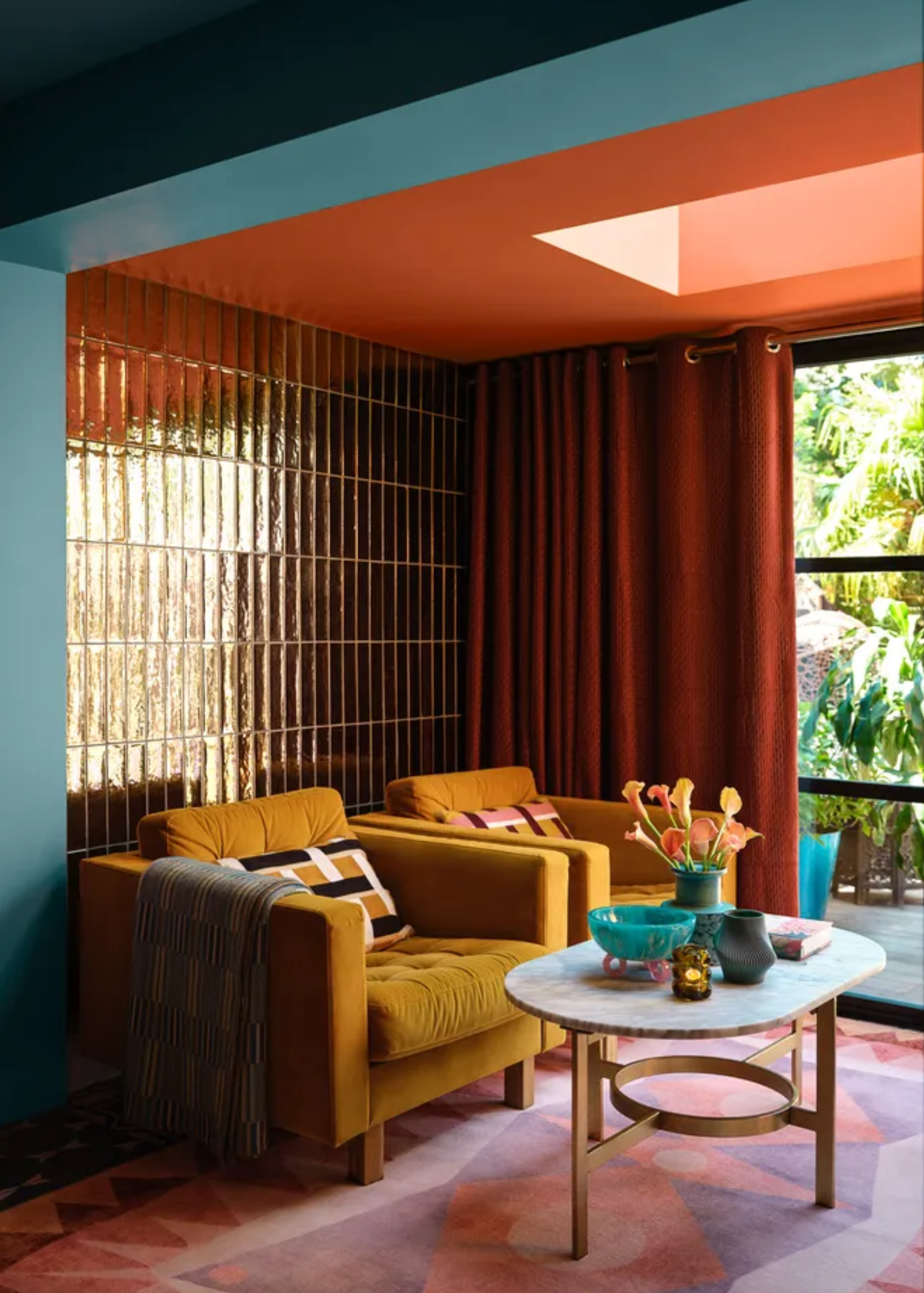 A living room with an orange ceiling and skylight, a shiny tiled accent wall, orange curtains, yellow plush armchairs, and a vase with flowers
