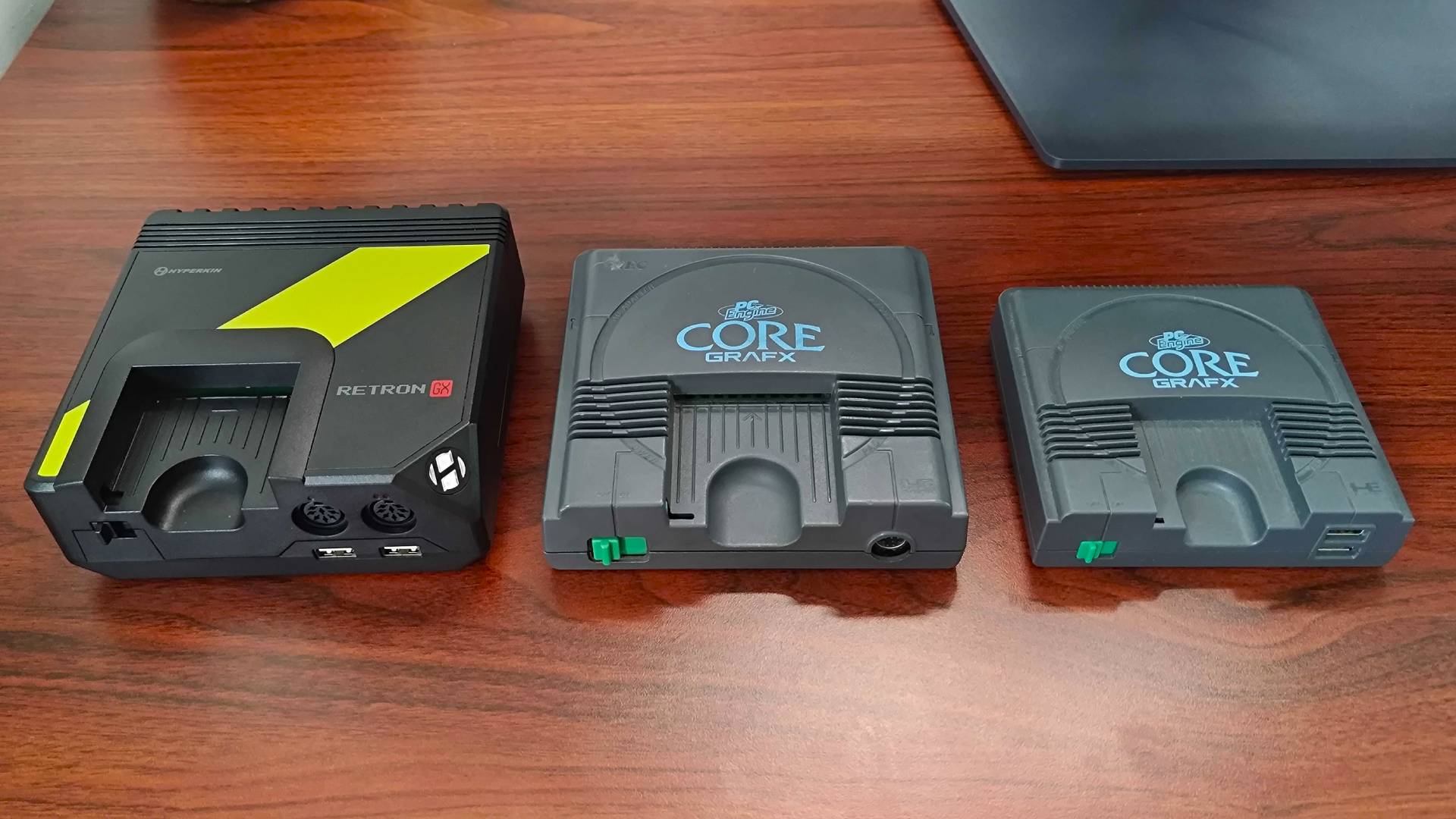 Hyperkin Retron GX on left with PC Egnine Core Grafx in centre, and PC Engine Mini on right sitting on woodgrain desk.