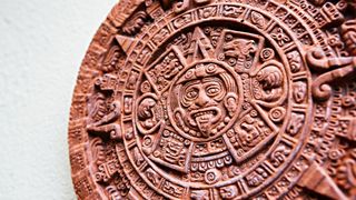 Here we see the Aztec calendar stone known as the Stone of the Sun. It is a round shape, with a face in the middle with it’s tongue out. In concentric circles going out from the middle there are several layers, each with intricate patterns.