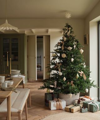 A realist christmas tree sparely decorated in a large pot surrounded by neutrally wrapped presents, in a neutrally decorated dining room