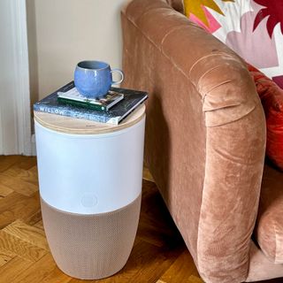 A Dupray Bloom air purifier as an accent table with books and a blue cup in a living room with orange loaf sofa and colourful cushions