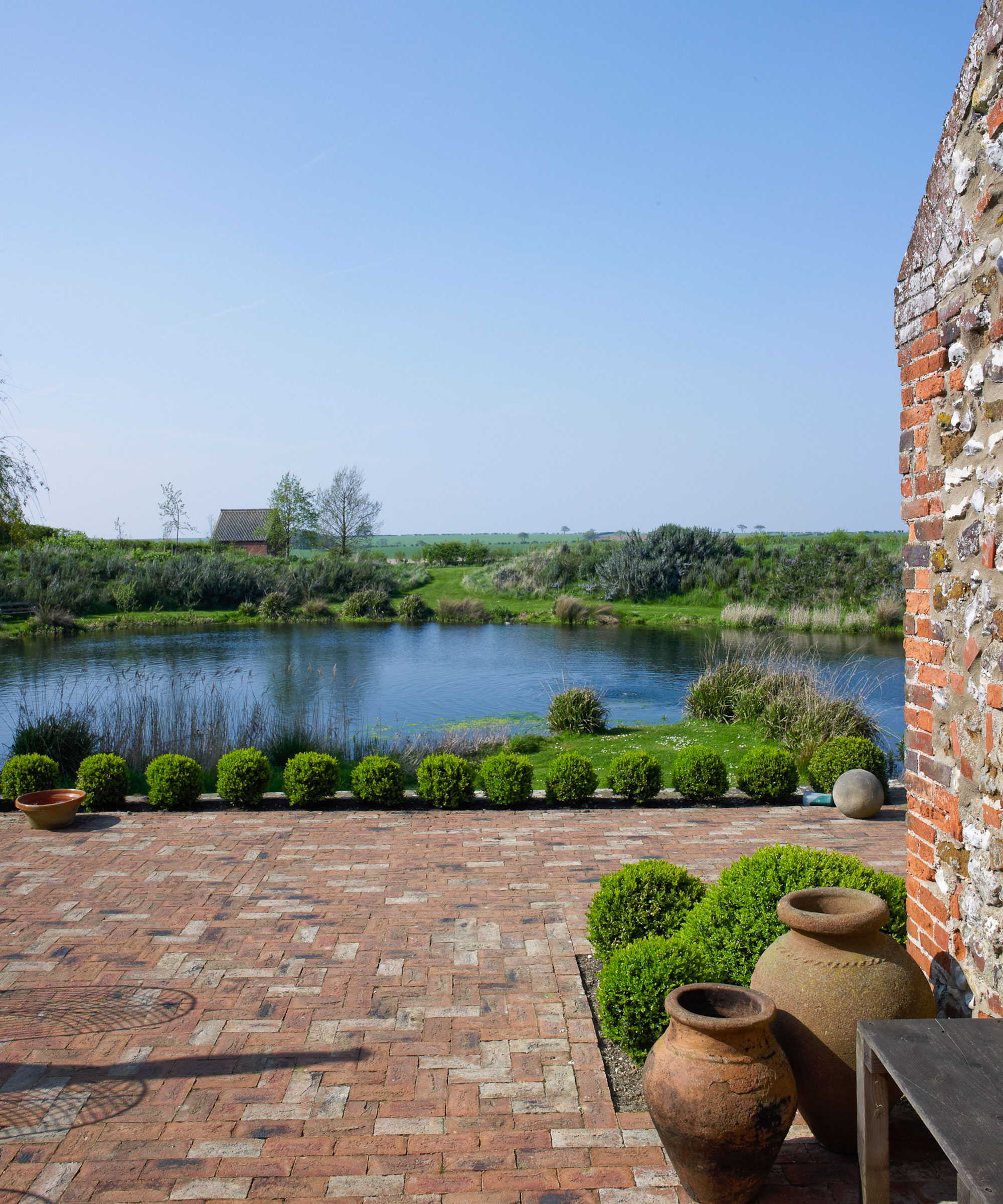 A brick paved courtyard with small round hedges looking onto a lake and fields.