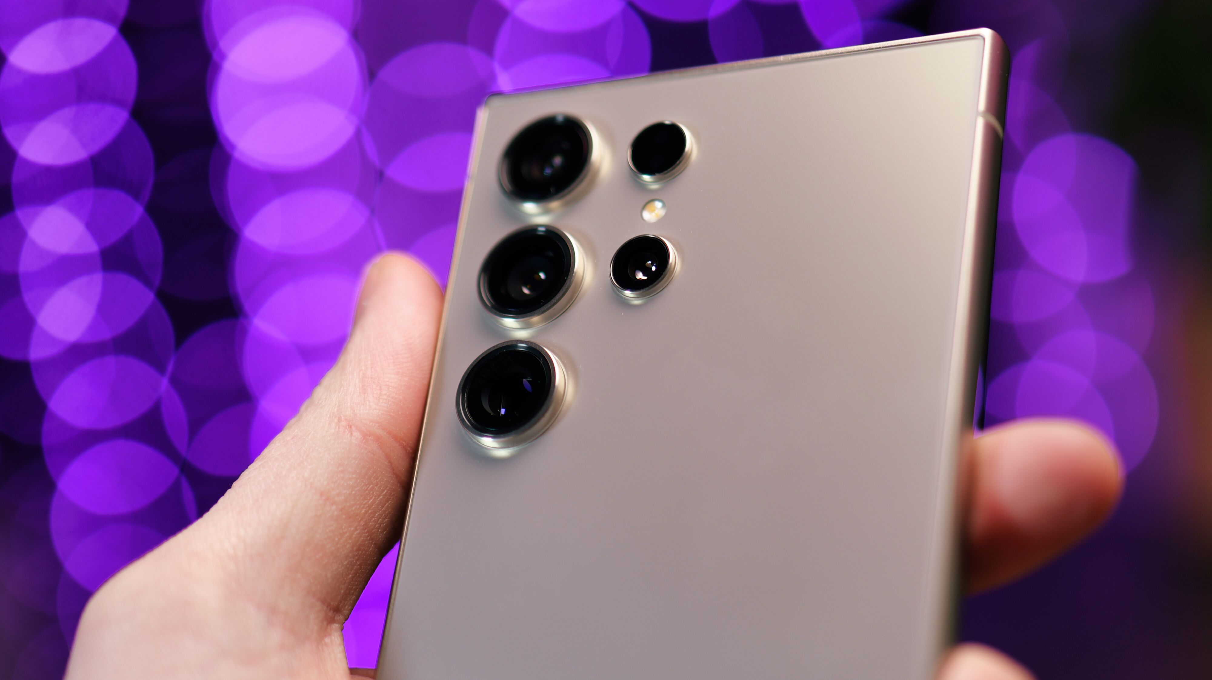 Gros plan des caméras d'un téléphone Samsung Galaxy S24 Ultra tenu dans une main avec des lumières violettes floues en arrière-plan