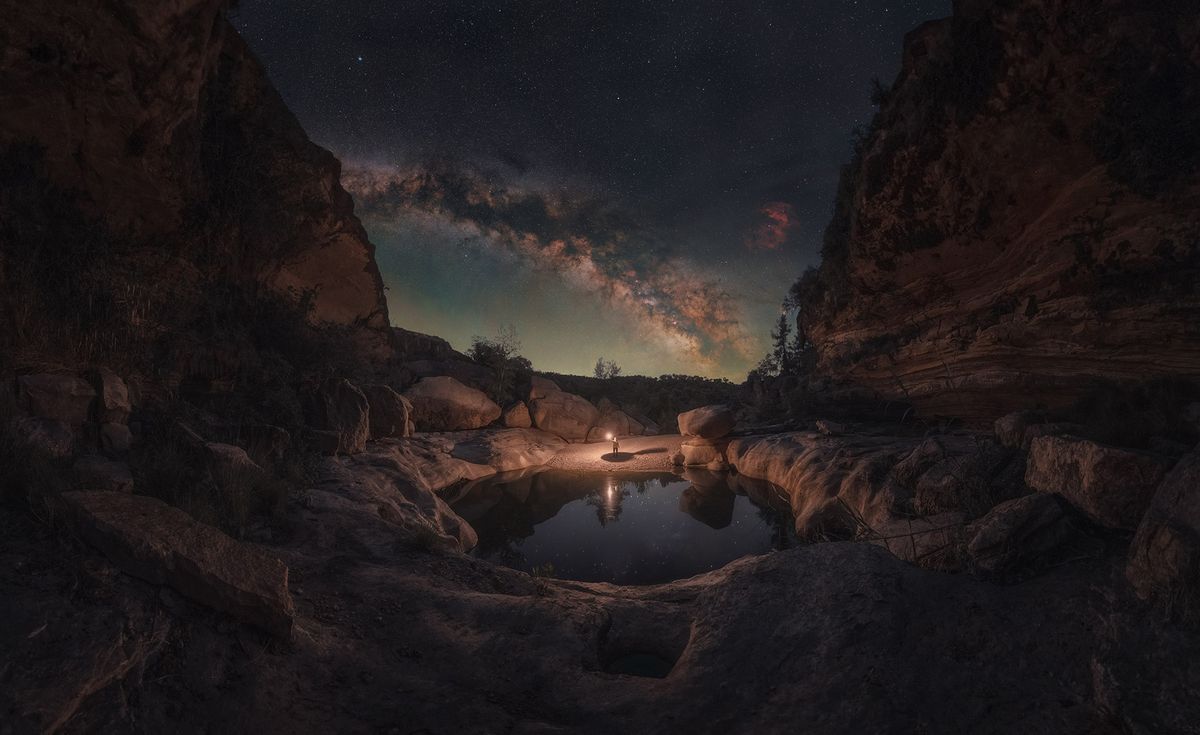 A person stands by a serene pond illuminated by a lantern under a starry sky, with the Milky Way arching overhead between rocky cliffs