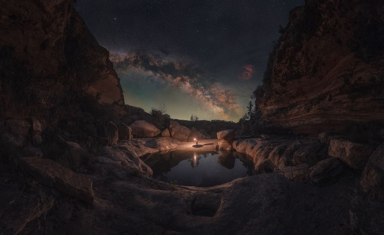 A person stands by a serene pond illuminated by a lantern under a starry sky, with the Milky Way arching overhead between rocky cliffs