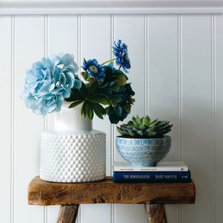 Blue flowers in white vase on wooden stool, white wall panelling, blue painted wall