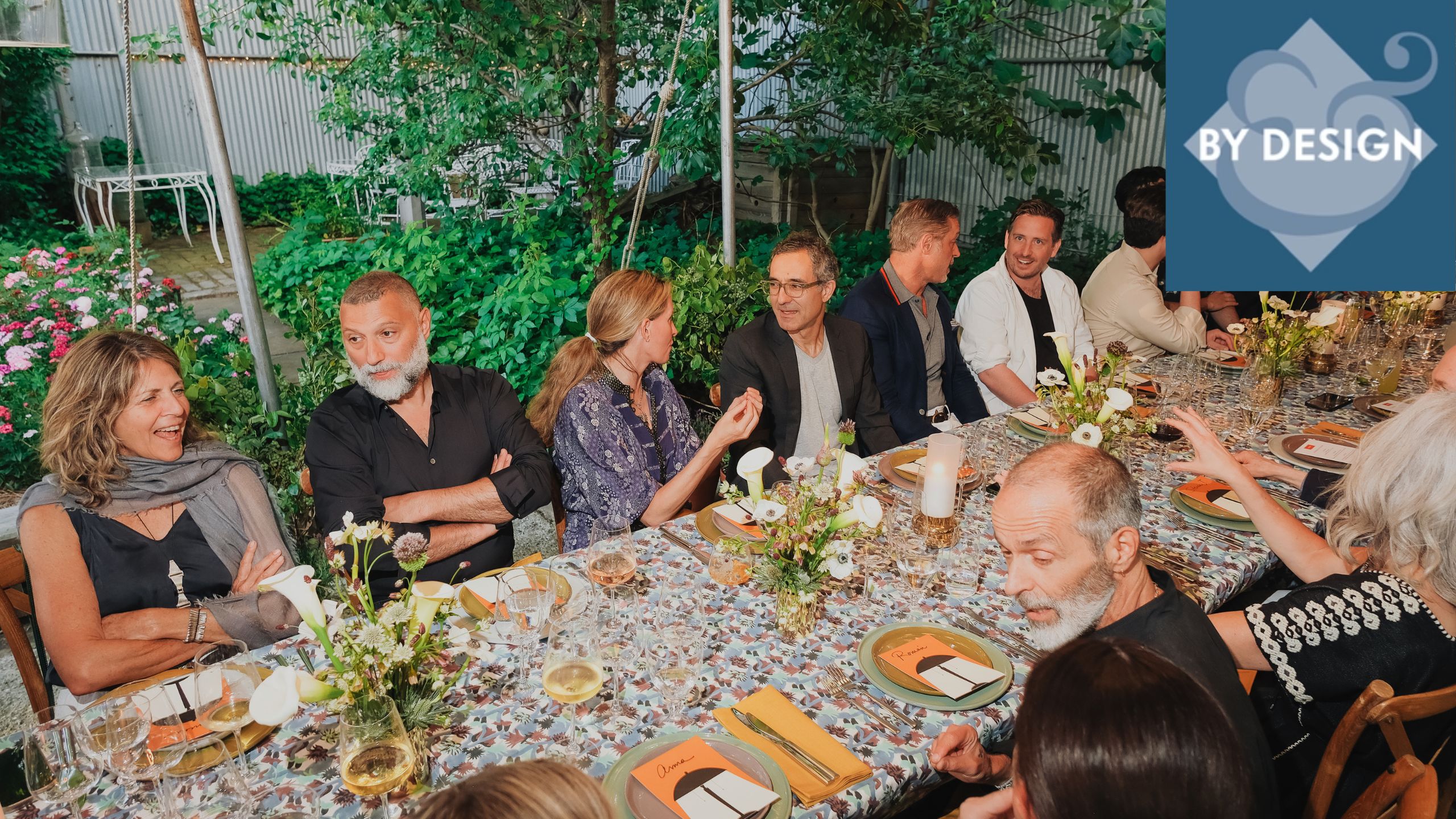 Photo of a large group of people sitting around a long rectangular dining table outdoors. Table covered in floral tableware and dinnerware, garden in the background
