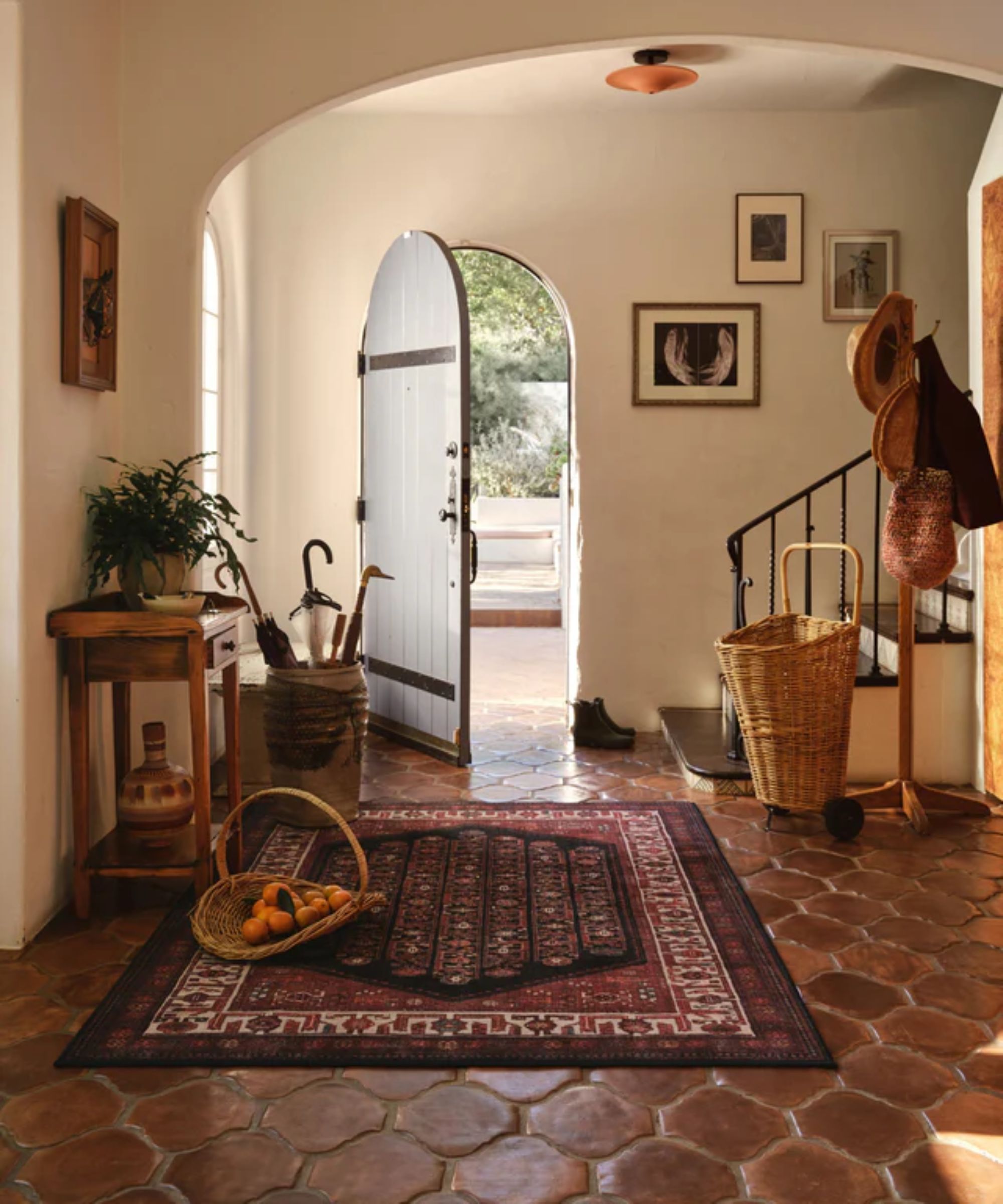 neutral entryway in a spanish style home with arched front door and dark terracotta floors styled with a large dark red rug, wooden table, and vintage baskets