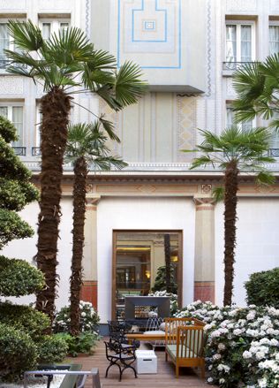 Prince de Galles, Paris, France. The outside of a hotel which has a seating area with tables and chairs surrounded by plants, flowers and palm trees.