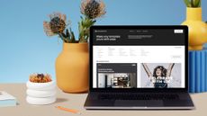 A laptop sitting on a desk displaying Squarespace on the screen, with a pen, two plants in pots, and a paper calendar 