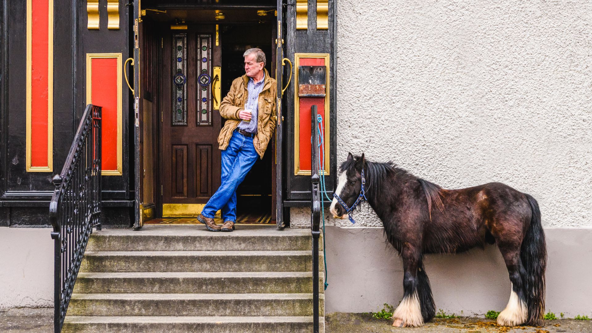 Black pony tied outside the entrance to a pub, steps lead up to the door where his owner stands