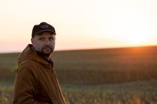 Outside image of Tomás Saraceno, brown hooded jacket and black baseball cap, blurred grass landscape, glow of low sun in a bright sky