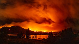 A forest fire burns behind a lake and its surrounding forest.