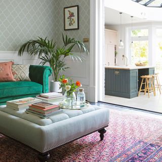 Wallpapered living room extending to kitchen, teal velvet sofa and ottoman coffee table with patterned rug