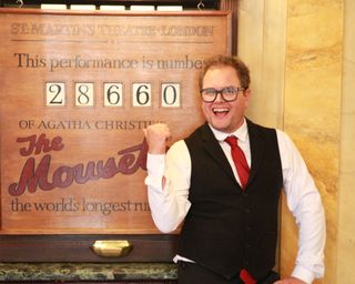 Alan Carr&#039;s adventures with Agatha Christie - Alan Carr stands in front of the sign at St Martin&#039;s Theatre reading &#039;This performance is number 28660 of Agatha Christie&#039;s The Mousetrap&#039;. He is dressed in a theatre front-of-house uniform of a white shirt, red tie, and a black waistcoat with matching trousers