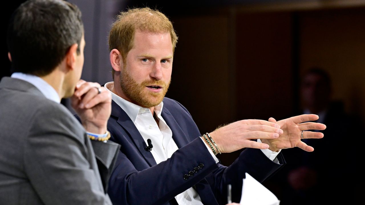 Prince Harry wearing a navy blazer and white shirt speaking to a man in a gray suit whose back is turned to the camera