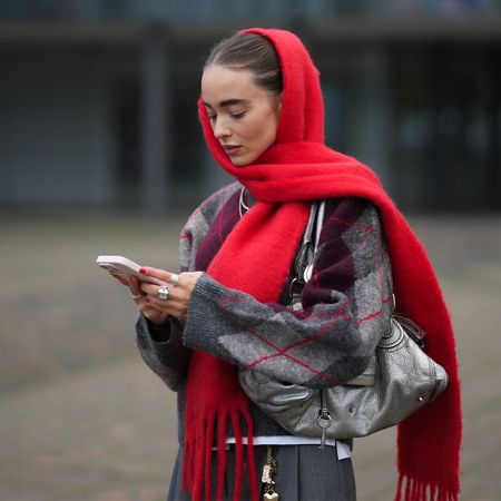 Woman in red knit scarf, argyle print sweater, silver metallic bag, and grey trousers, on her phone. 