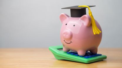 Piggy bank wearing a mortarboard graduation cap standing on top of a green calculator on a wooden table