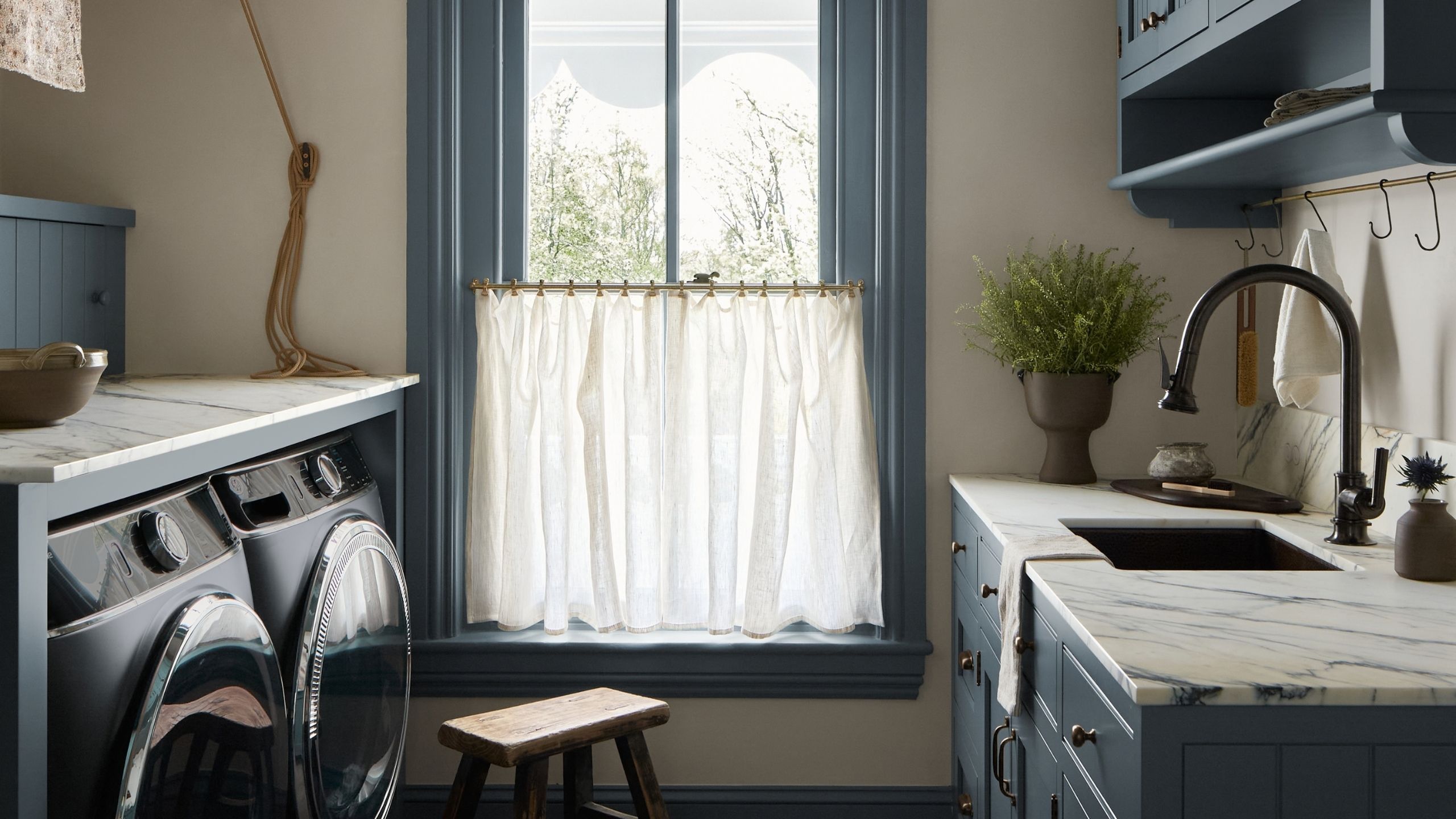 Blue and white laundry room with marble countertops and large window with cafe curtains